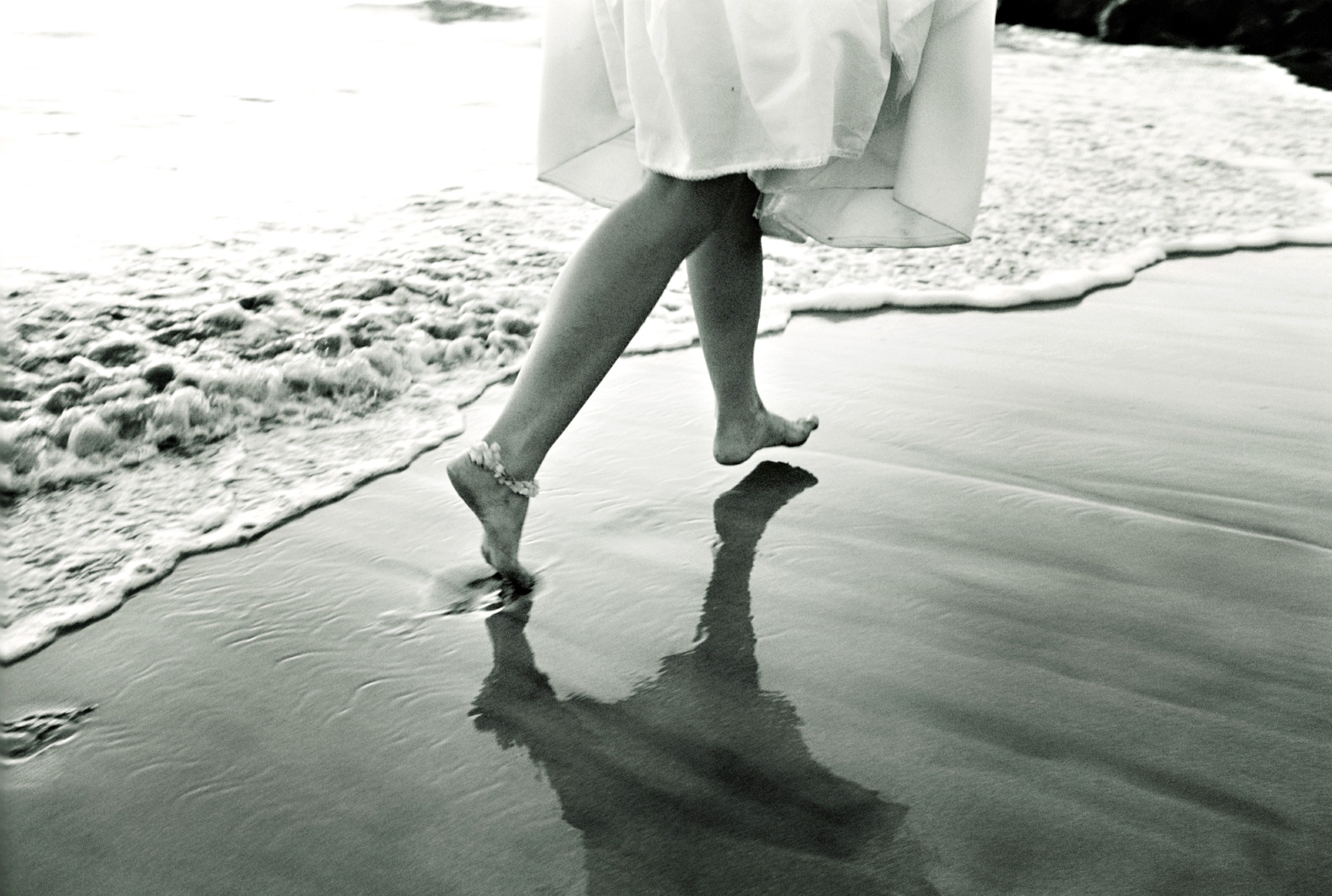 Black-and-white photograph of a barefoot bride stepping along the shoreline, her reflection falling across the wet sand