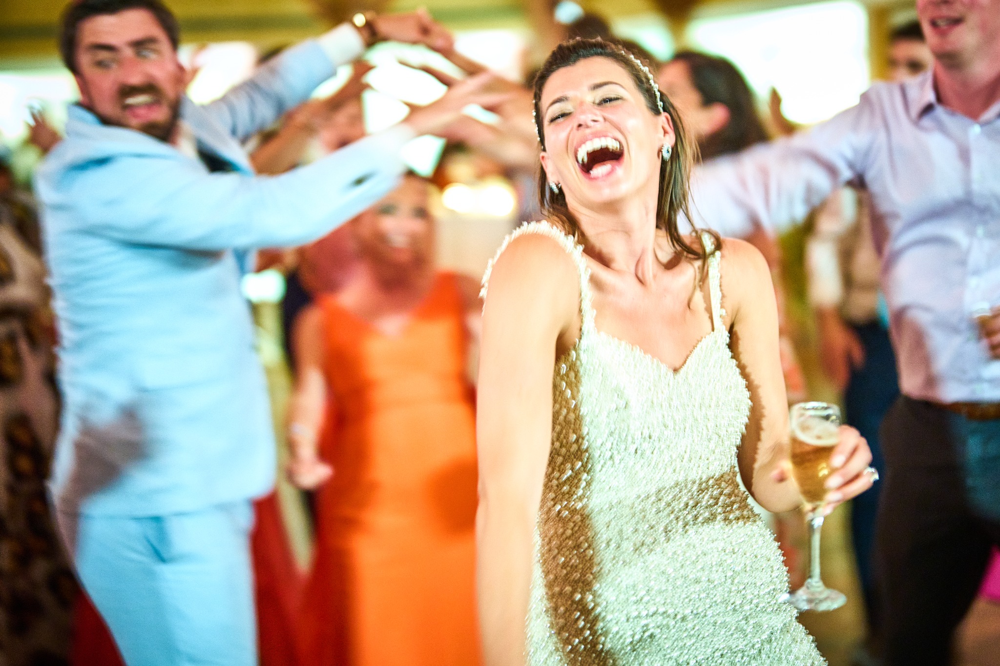 A laughing bride holding a champagne glass amid guests on the dance floor