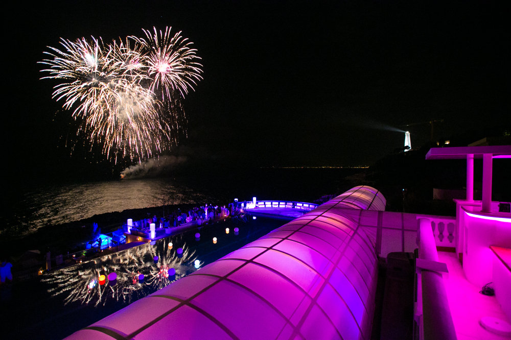 Fireworks over the water beside a wedding celebration illuminated in purple and blue