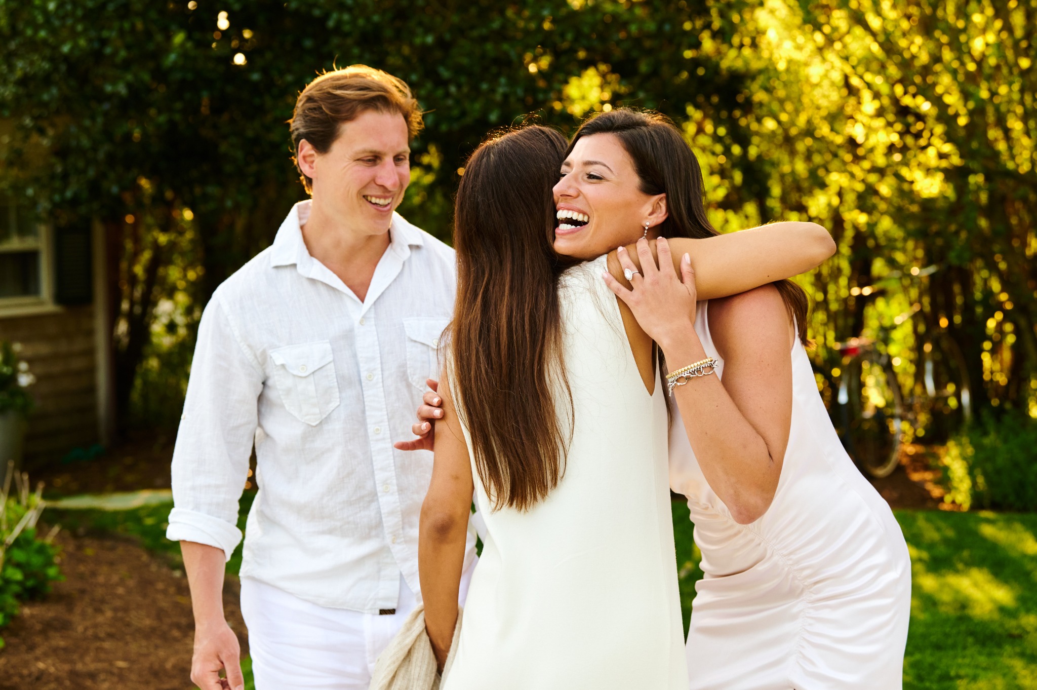 A smiling woman embracing a guest as her partner looks on at their outdoor wedding welcome party
