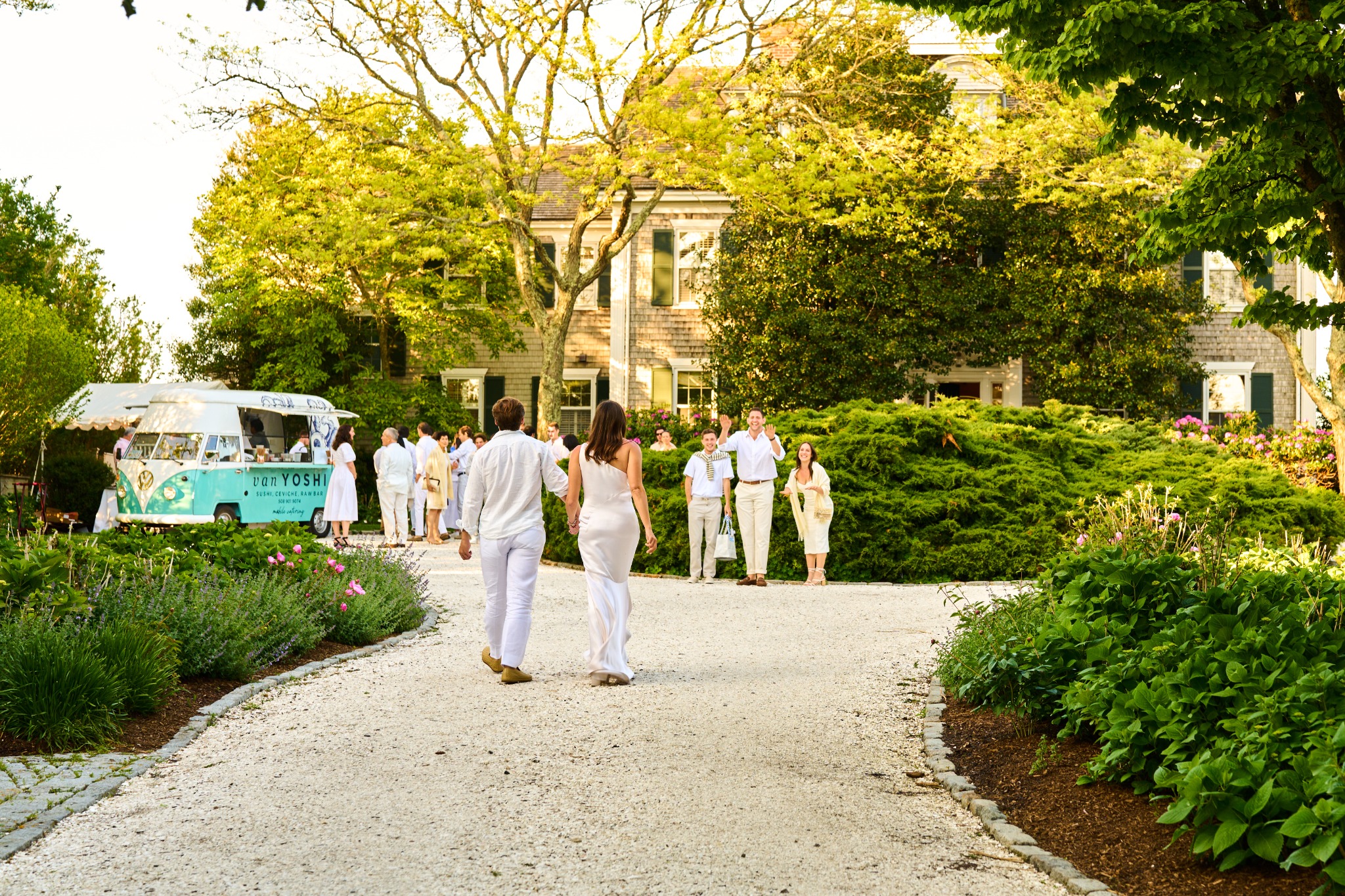 A couple walking hand in hand toward guests waving at an outdoor wedding welcome party